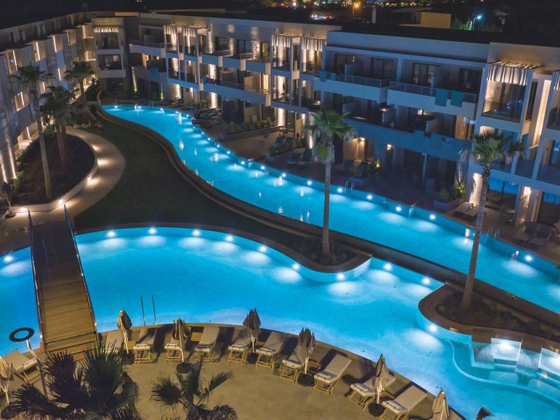 Piscine d'hôtel éclairée la nuit avec chaises longues et bâtiments modernes alentour.