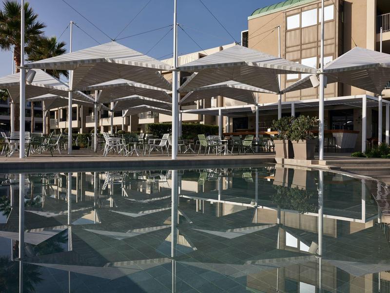 Piscine d'hôtel moderne avec parasols blancs et palmiers éclairés par le soleil.