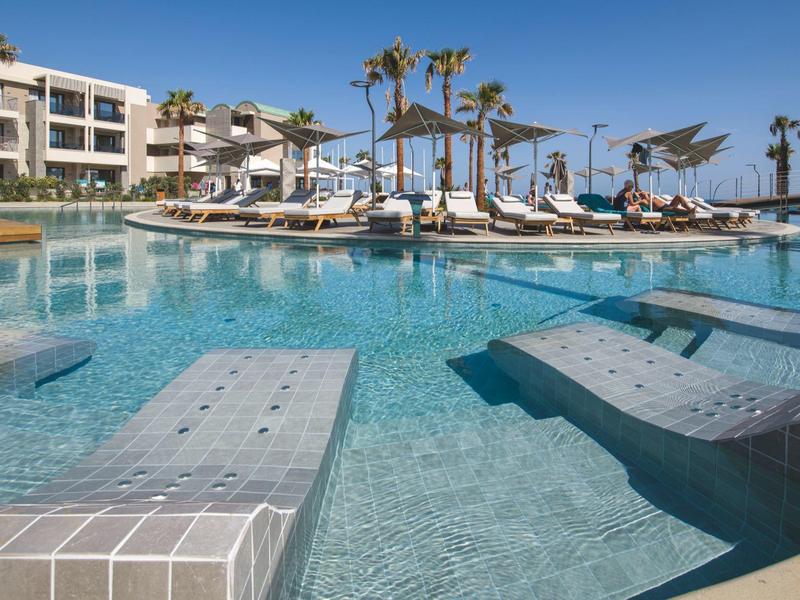 Piscine d'hôtel moderne avec transats intégrés, entourée de palmiers et de bains de soleil.