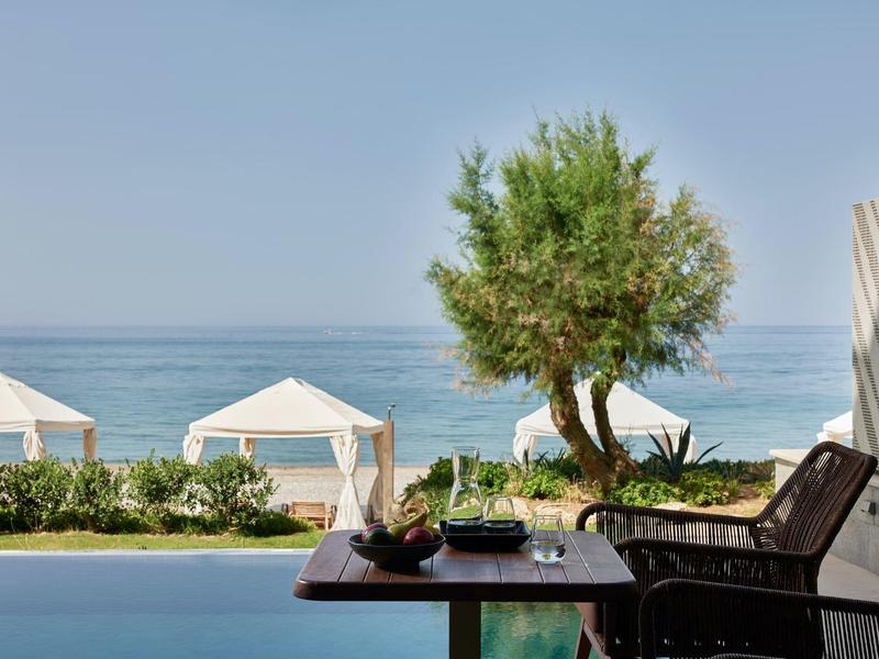 Terrasse avec table, chaises, olivier, vue sur la mer et cabanes blanches.