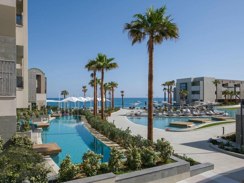 Espace piscine moderne d'hôtel avec palmiers et vue sur la mer sous un ciel clair.