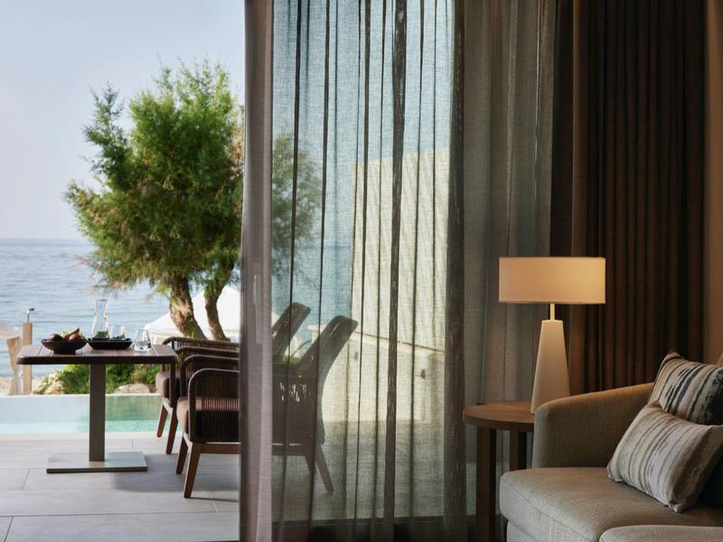 Vue à travers des rideaux transparents vers une terrasse avec table et olivier au bord de la mer.