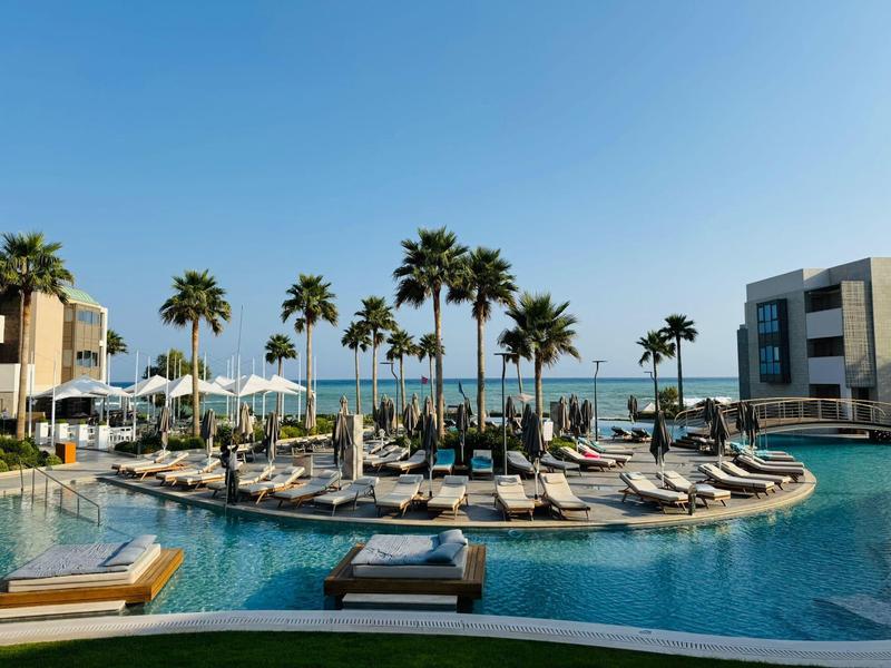 Piscine d'hôtel avec chaises longues, palmiers et vue sur la mer sous un ciel clair.