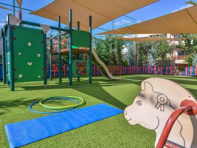 Bunter Kinderspielplatz mit Kletterwand, Rutsche, Schaukelpferd und Matte auf grünem Kunstrasen.