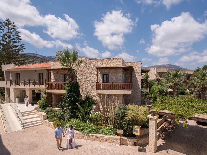 Grande villa en pierre avec terrasses entourée de végétation luxuriante sous un ciel partiellement nuageux.