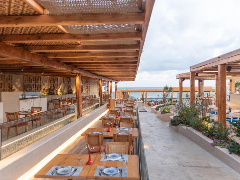 Terrasse de restaurant extérieure avec tables en bois et vue sur la mer sous des pergolas en bois.