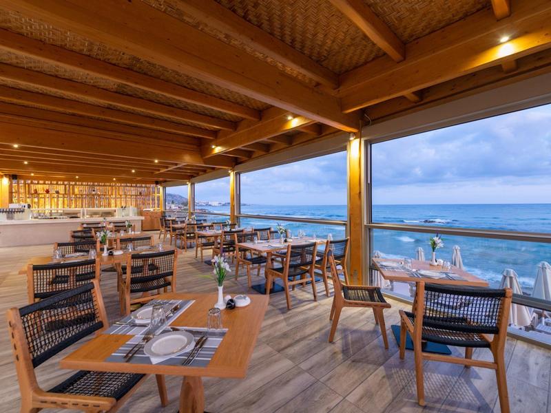 Restaurant en bord de mer avec tables et chaises en bois sous une terrasse couverte au coucher du soleil.