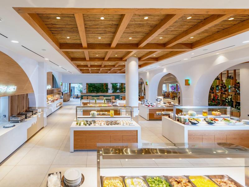 Buffet d'hôtel lumineux avec plafond en bois, diverses présentations de nourriture et décor blanc.