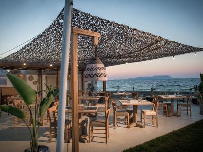 Terrassenbereich mit Tischen und Stühlen, überdacht von einem Netz, Meer im Hintergrund bei Sonnenuntergang.
