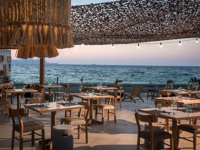 Offenes Strandrestaurant mit Holzstühlen, Tischen, Lampen und Blick auf das ruhige Meer.