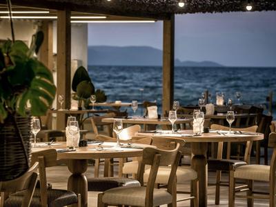 Restaurant mit Holztischen und Stühlen am Meer mit Blick auf ruhige Wellen und Berge im Hintergrund.
