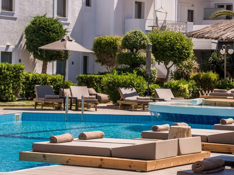 Reposeras modernas con cojines junto a una piscina azul clara, rodeadas de plantas verdes y edificios blancos.