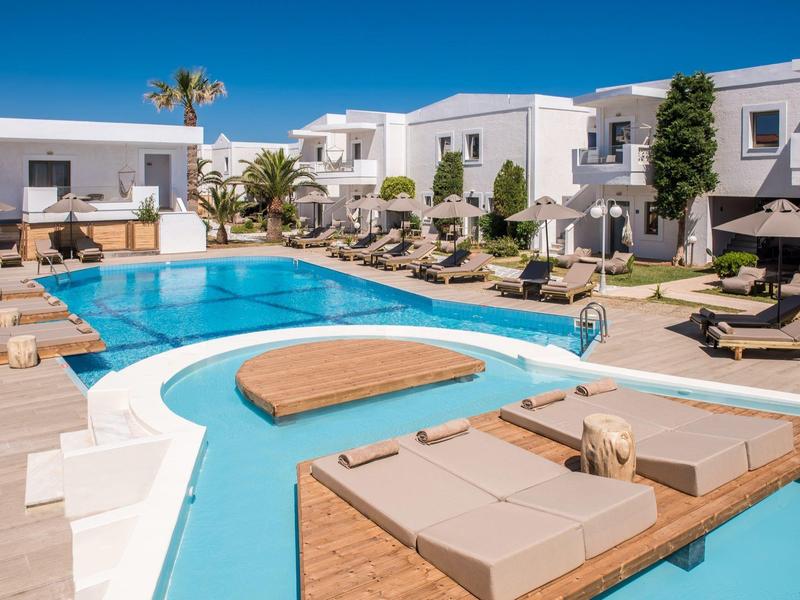 Área de piscina de hotel moderno con edificios blancos, tumbonas y sombrillas bajo un cielo azul.