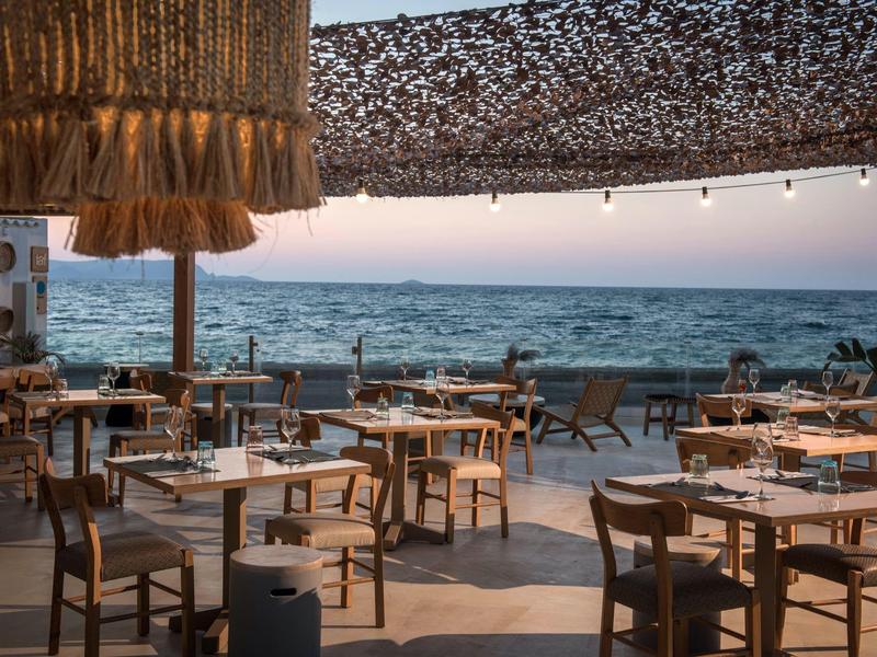 Restaurante al aire libre con muebles de madera y vista al mar al atardecer.