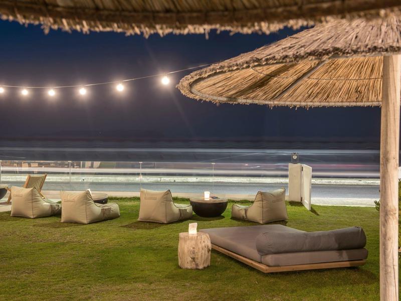 Área al aire libre frente a la playa con tumbonas, cojines y luces colgantes por la noche.