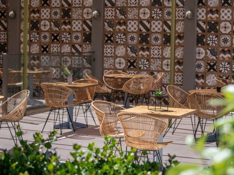 Área de terraza vacía con sillas y mesas de mimbre frente a una pared decorativa.