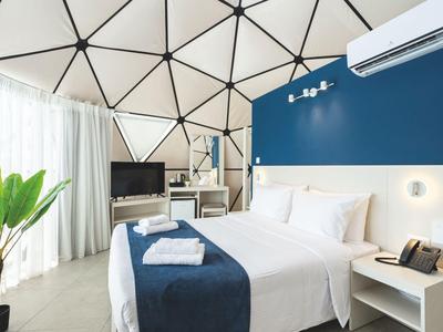 Moderní hotelový pokoj s manželskou postelí, modrou stěnou, televizí a oknem s bílou záclonou.