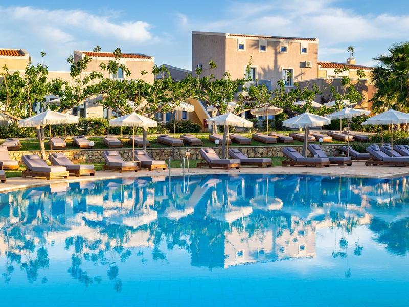 Duży basen z leżakami i parasolami przed hotelem pod niebieskim niebem.