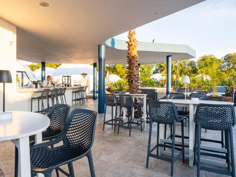 Nowoczesny zewnętrzny bar hotelowy z stołami, krzesłami i widokiem na palmy i wodę.