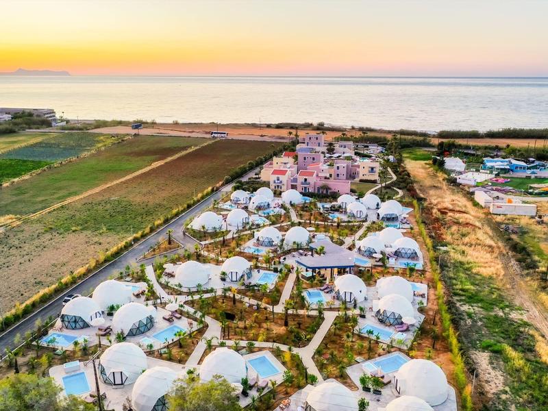Widok z powietrza na ośrodek z białymi namiotami kopułowymi obok zielonych pól i morza o zachodzie słońca.