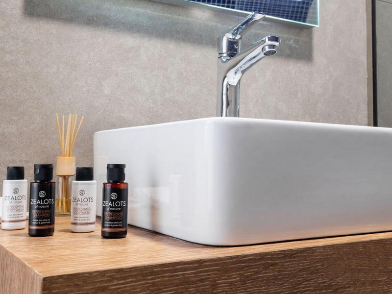 Biała umywalka na drewnianym blacie z butelkami kosmetycznymi i dozownikiem w łazience.