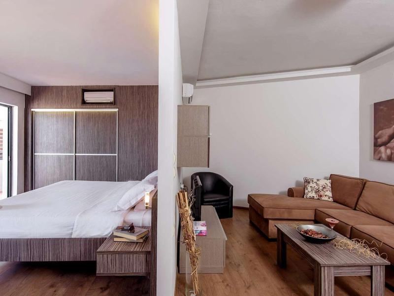 Helles Schlafzimmer mit Balkon und angrenzendem Wohnzimmer mit braunem Sofa und Tisch.