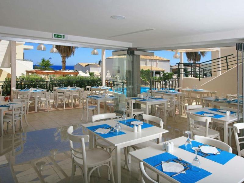 Helles Restaurant mit weißen Tischen und blauen Tischsets, Blick auf Terrasse und Palmen.