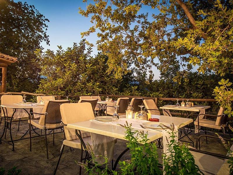 Freiluft-Café mit weißen Tischen und schwarzen Stühlen umgeben von grünen Bäumen im Sonnenlicht.