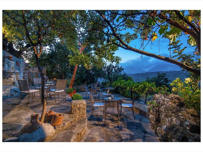 Terrasse mit Stühlen und Tischen unter Bäumen, Blick auf Berge und blauen Himmel.