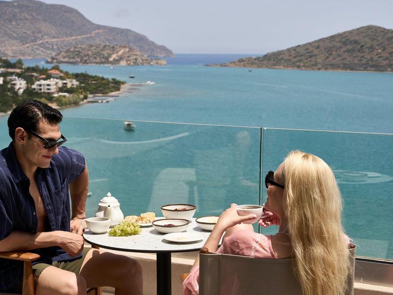 Dos personas sentadas en una terraza con vistas al mar y montañas en un día despejado.
