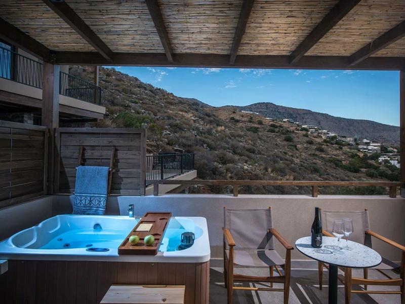 Balcón con jacuzzi, dos sillas y mesa con botella de vino, vista a paisaje montañoso.