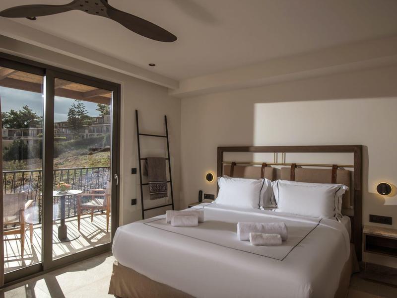 Světle zařízený hotelový pokoj s velkou postelí, balkonem a výhledem na město.