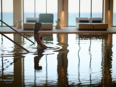 Woman steps into a heated indoor pool with sea view and modern furnishings.