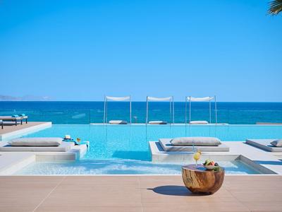 Luxury pool area with sea view, sun loungers, and canopy beds under a blue sky.