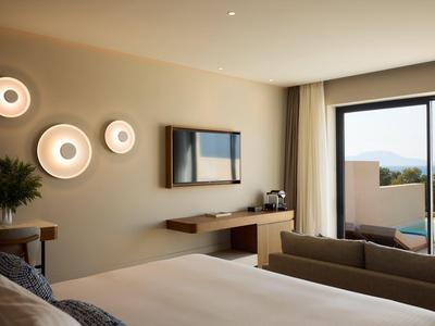 Moderne hotelkamer met groot bed, bank, muurverlichting en uitzicht op zwembad en zee.