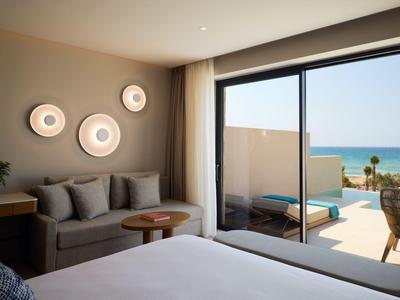 Bright hotel room with double bed, sofa, wall lights, and sea view through large glass windows.