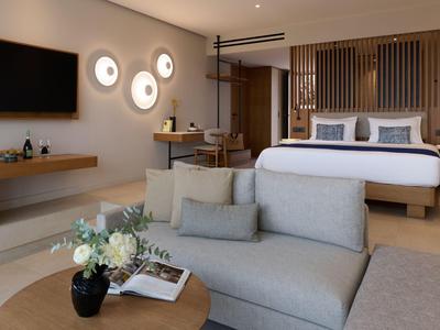 Modern hotel room with double bed, sofa, desk, and TV in warm tones.