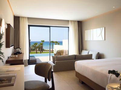 Bright hotel room with double bed, seating area, and view of pool and sea.