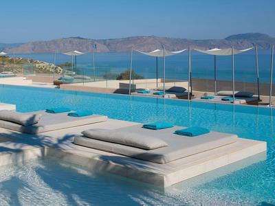 Modern infinity pool with loungers overlooking the sea and mountains in the background.