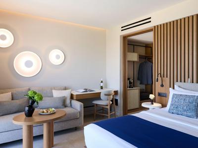 Modern hotel room with bed, sofa, small table, and workspace in neutral tones.