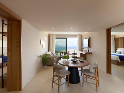 Modern hotel room area with dining table, chairs, sofa, and sea view through large windows.