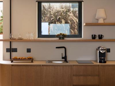 Modern kitchen area with wooden cabinets, a black faucet, and shelves in front of a window.