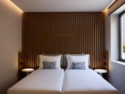 Modern hotel room with two single beds, wooden wall, and window letting in daylight.
