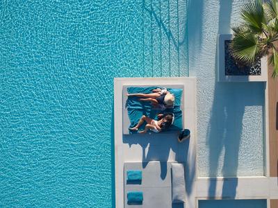 Two people lying on blue loungers in a clear pool with palm tree shadows.