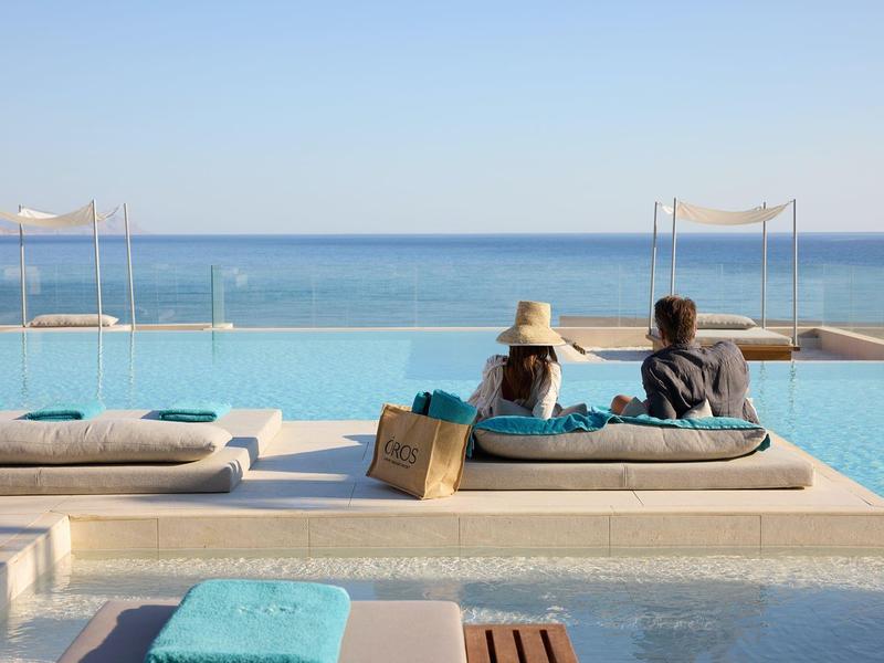 Two people relax at the infinity pool overlooking the sea under a clear sky.