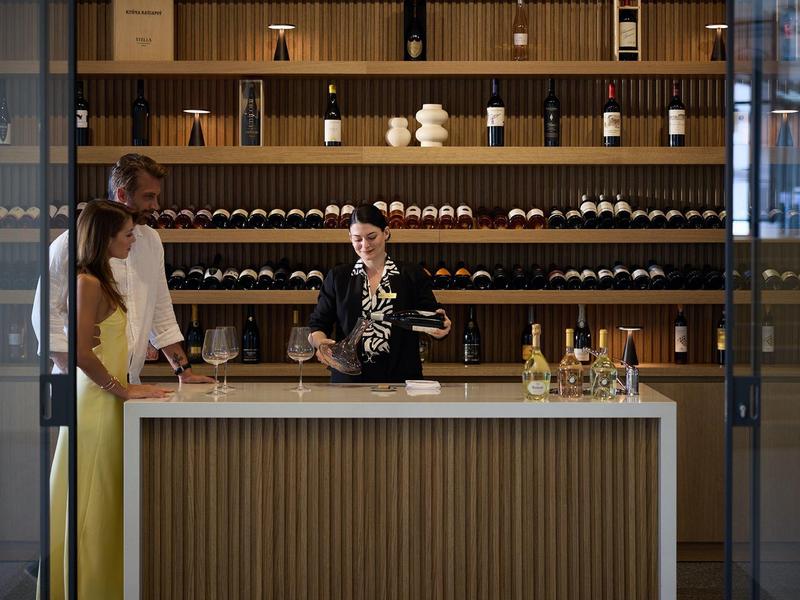 Hotel bar with bartender mixing cocktails and guests in front of a wine bottle shelf.