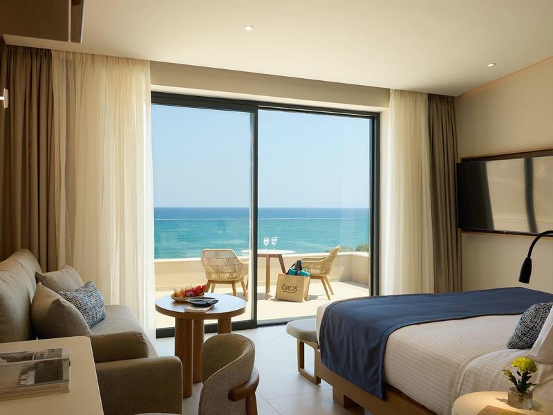 Modern hotel room with bed, seating area, and sea view through large windows.