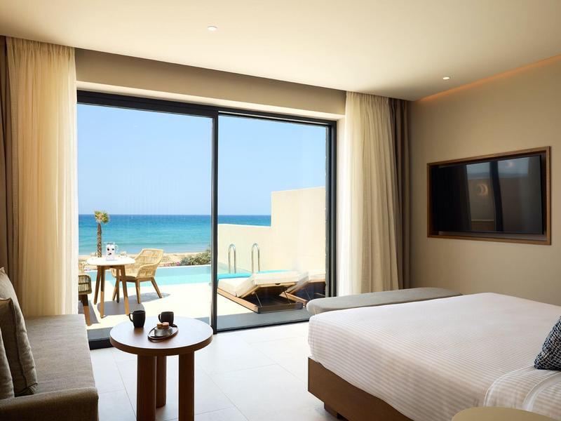 Bright hotel room with sea view, large bed, sofa, table, and terrace with chairs.