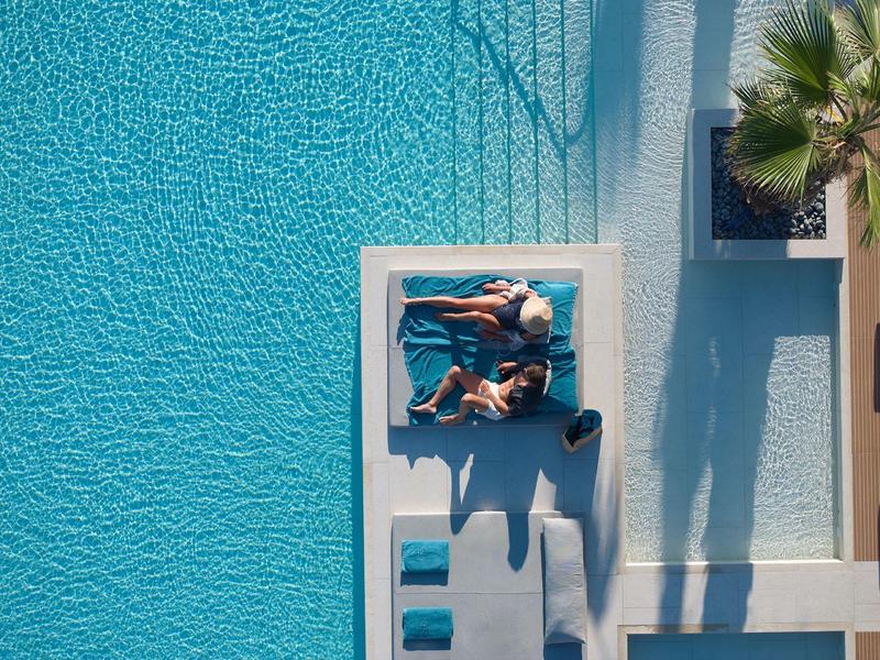 Two people relaxing on loungers by a pool with blue water and palm tree shadows.