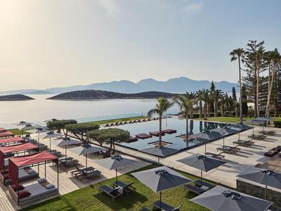 Moderner Poolbereich mit Liegestühlen und Pavillons vor einer Meeresbucht und Bergen.